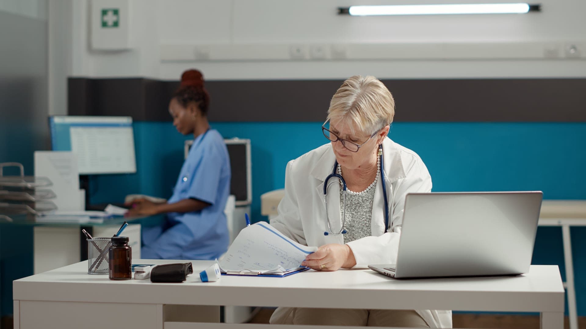 Médecin dépouillant une pile de dossiers à son bureau, secrétariat médical en arrière-plan
